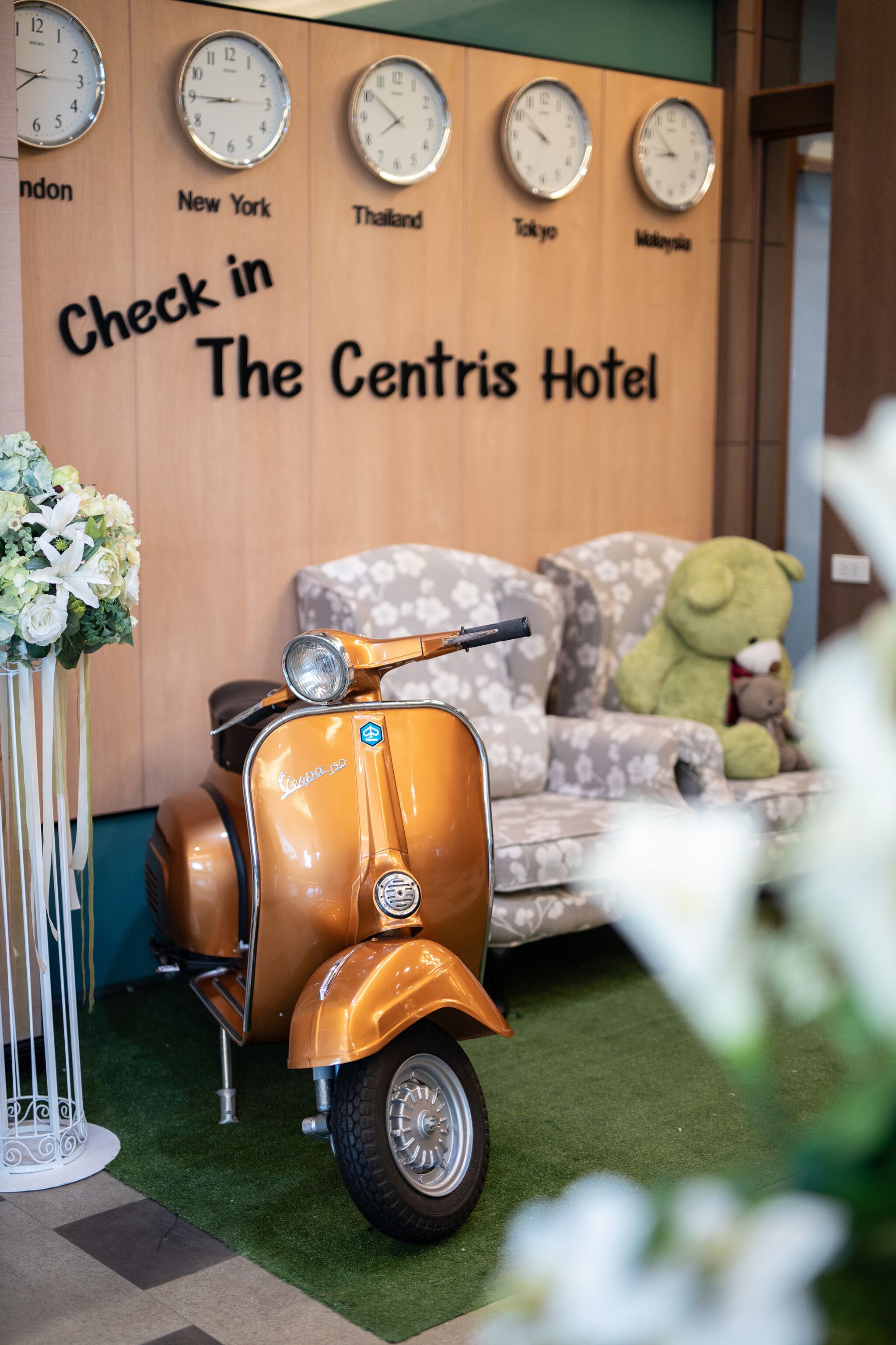 A vintage orange Vespa scooter, a plush sofa with a teddy bear, and a wall with clocks indicating different cities.