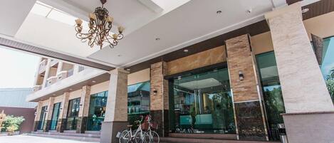 Entrada de hotel com design moderno, apresentando um candelabro, grandes janelas de vidro e uma bicicleta estacionada na parte externa.