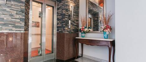 Un vestíbulo moderno con una pared de piedra, un ascensor de vidrio, una mesa consola de madera con un jarrón de flores y un espejo grande.