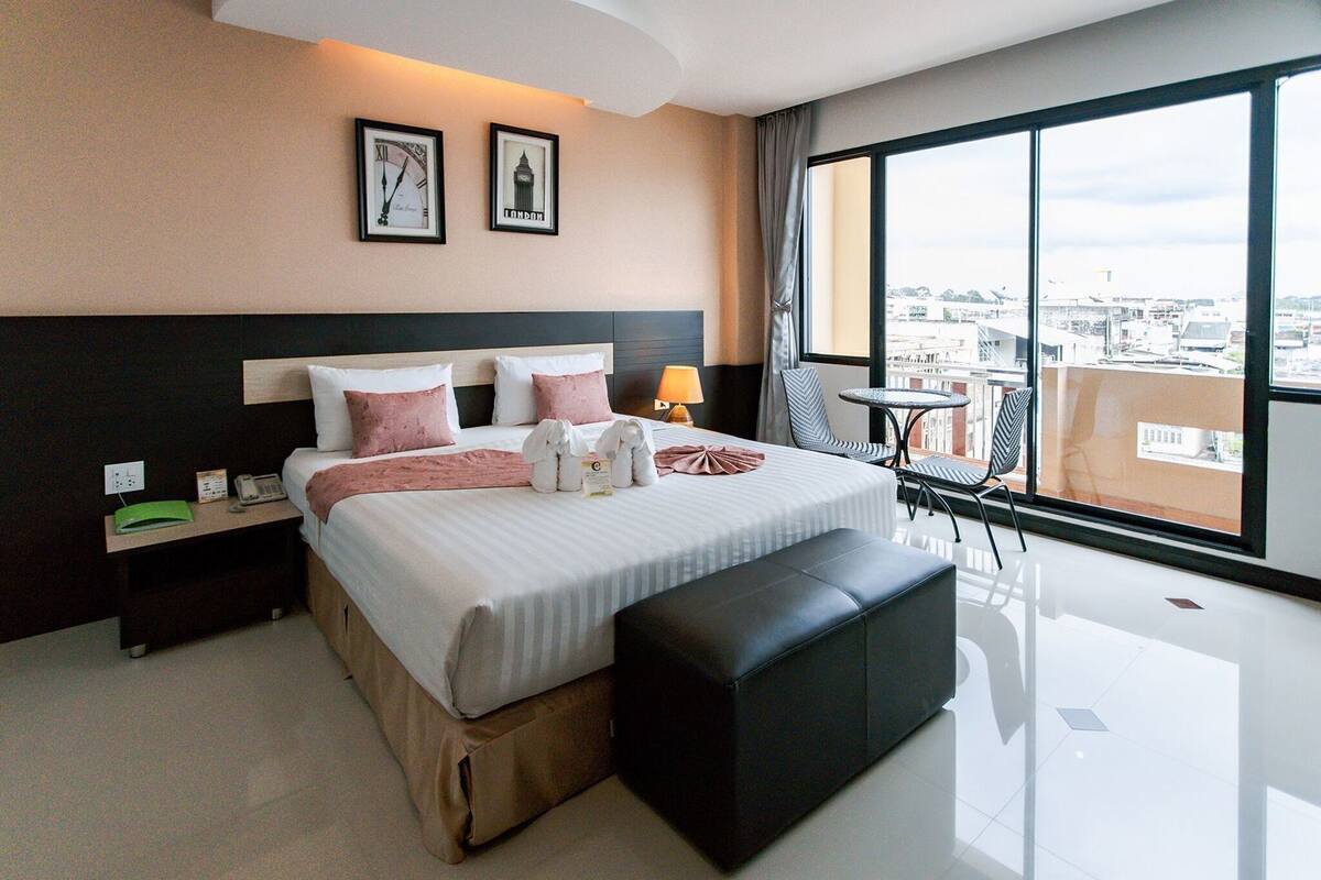 Habitación de hotel con cama, mesita de noche, lámpara y vistas a la ciudad a través de amplios ventanales.