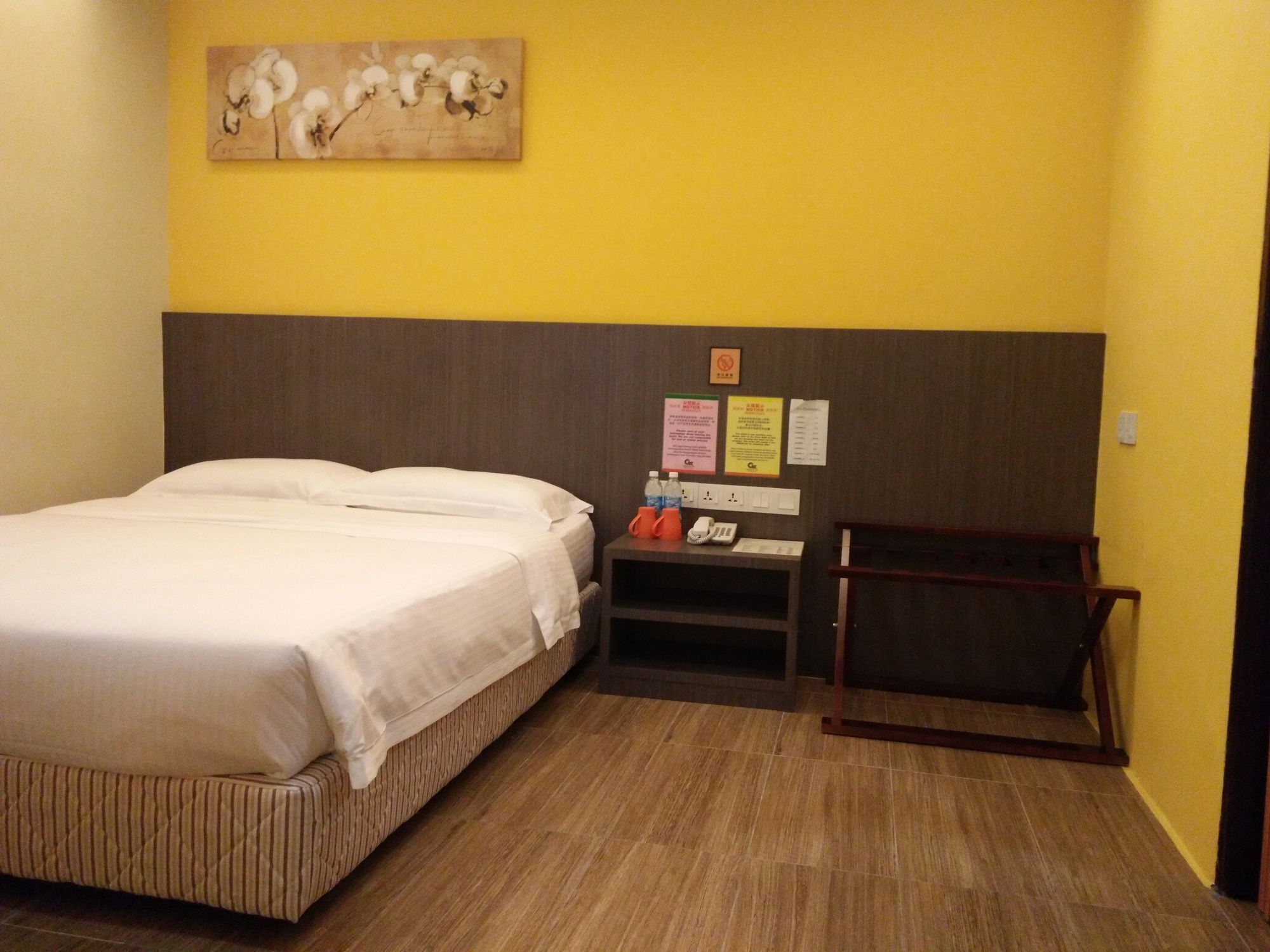 A hotel room with a bed, a nightstand, a wooden headboard, a wall-mounted artwork, and a yellow wall.