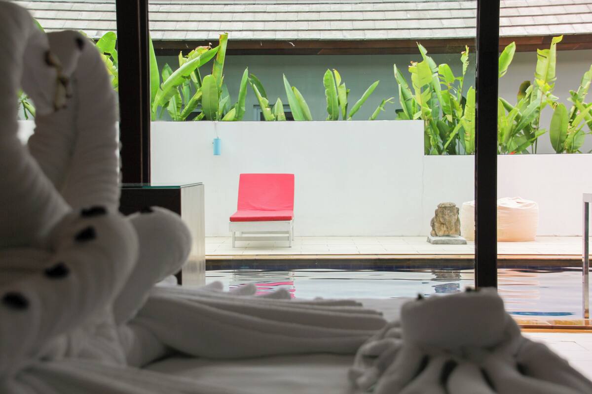 A hotel room with a view of a pool, a red lounge chair, and green plants.