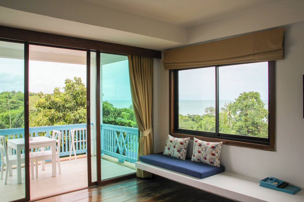 A room with a balcony, a seating area with cushions, and a view of the outdoors.