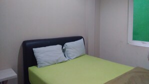 Ein Einzelbett mit einer grünen Matratze und zwei weißen Kissen, ein weißer Nachttisch und ein Fenster mit einer grünen Plane.