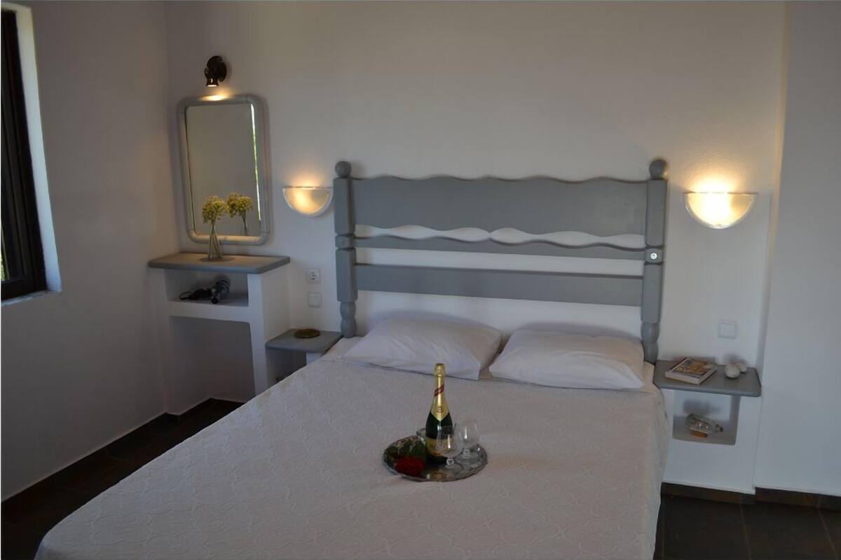 A neatly made bed with a bottle of champagne, a bedside table with a lamp, and a nightstand with a lamp.