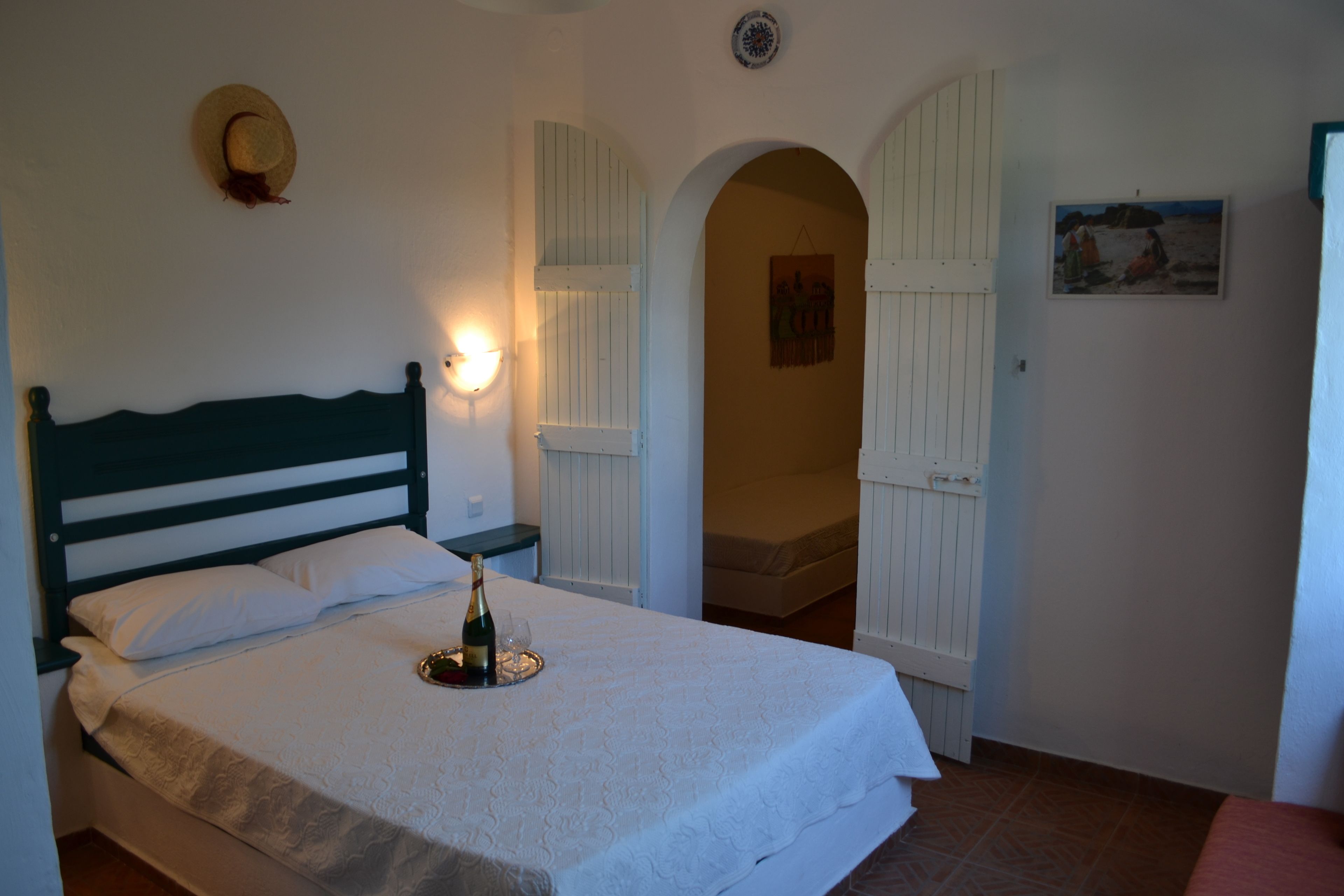A bedroom with a bed, a bedside table with a bottle and glass, a wall-mounted hat, and an archway leading to another room.