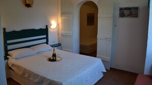 A bedroom with a bed, a bedside table with a bottle and glass, a wall-mounted hat, and an archway leading to another room. - Kochylas Studios (Skiros)
