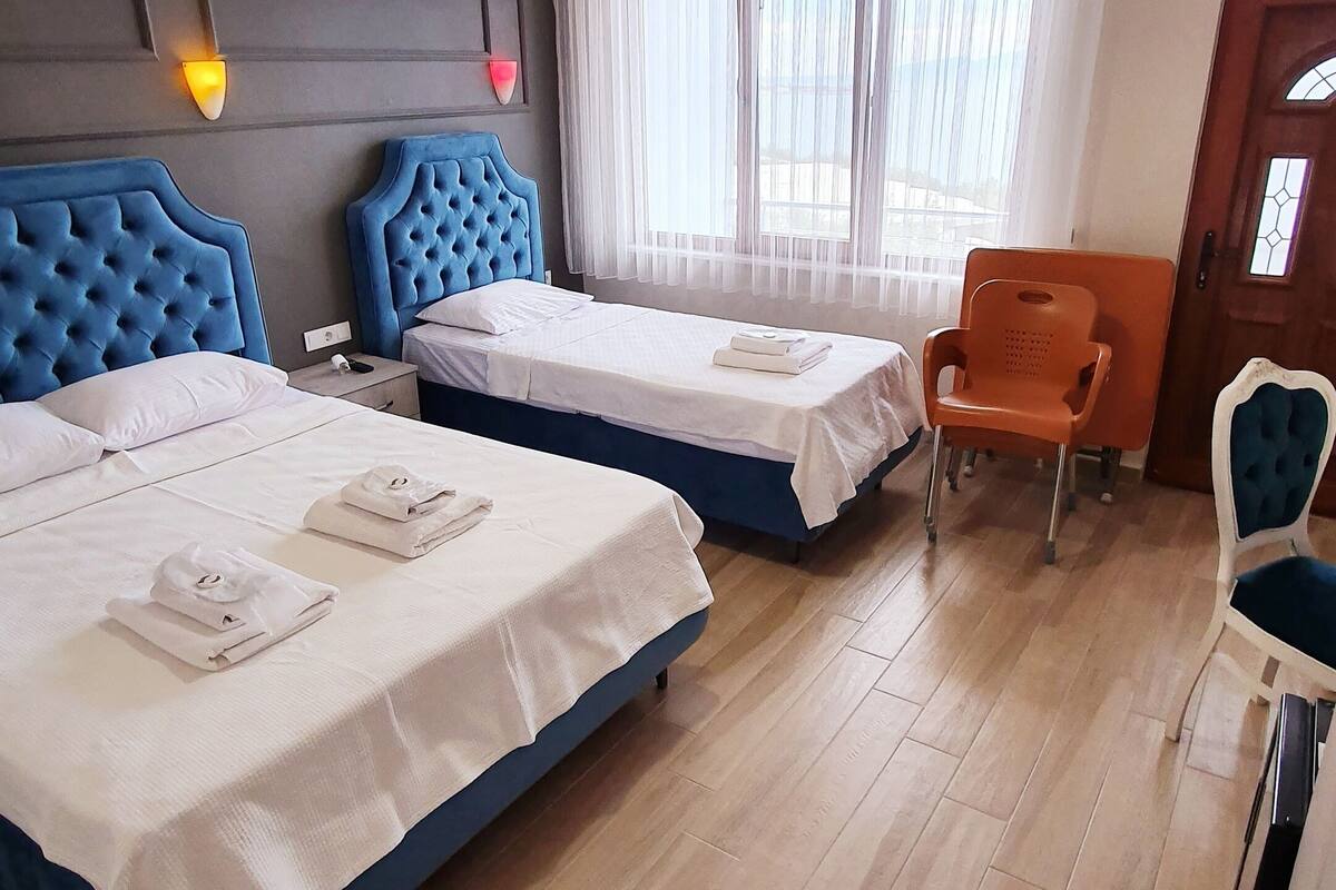 A hotel room with two beds, a wooden floor, a large window with curtains, a wooden door, and a chair.