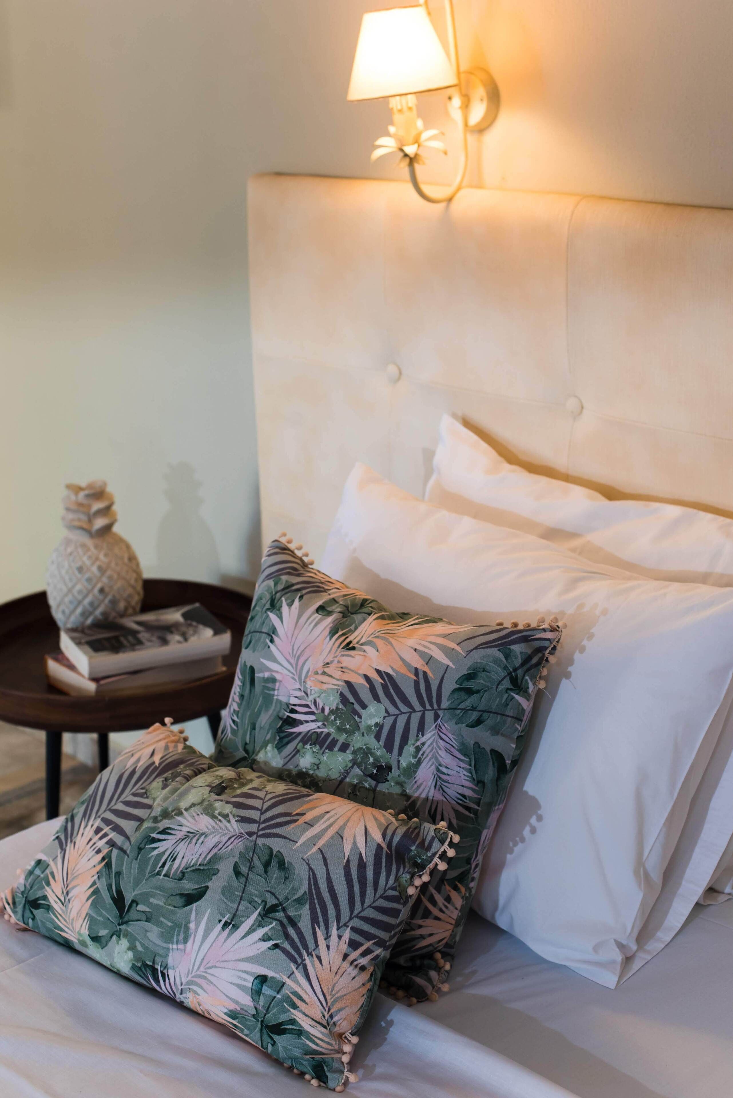 Una cama con sábanas blancas, dos almohadas de estampado floral, una mesita redonda con un adorno y una lámpara de pared.