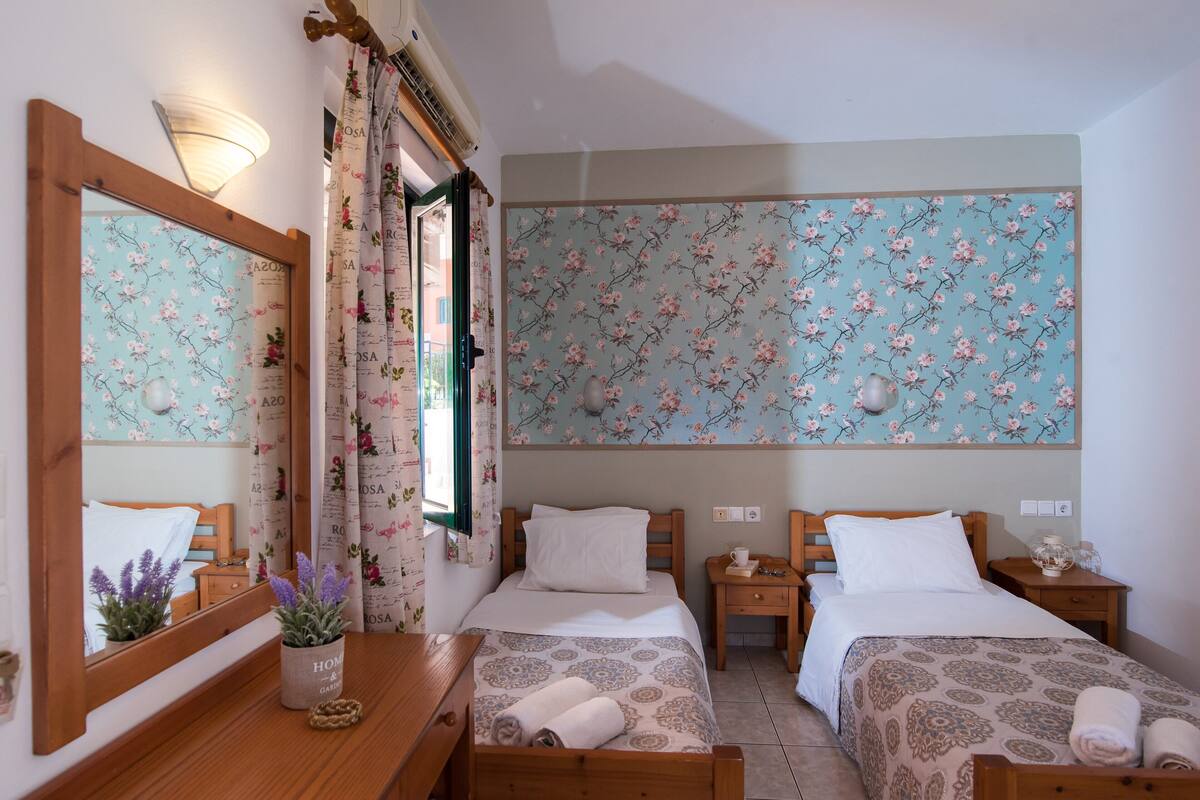 A hotel room with two single beds, a wooden dresser, a mirror, and a floral-patterned wall.
