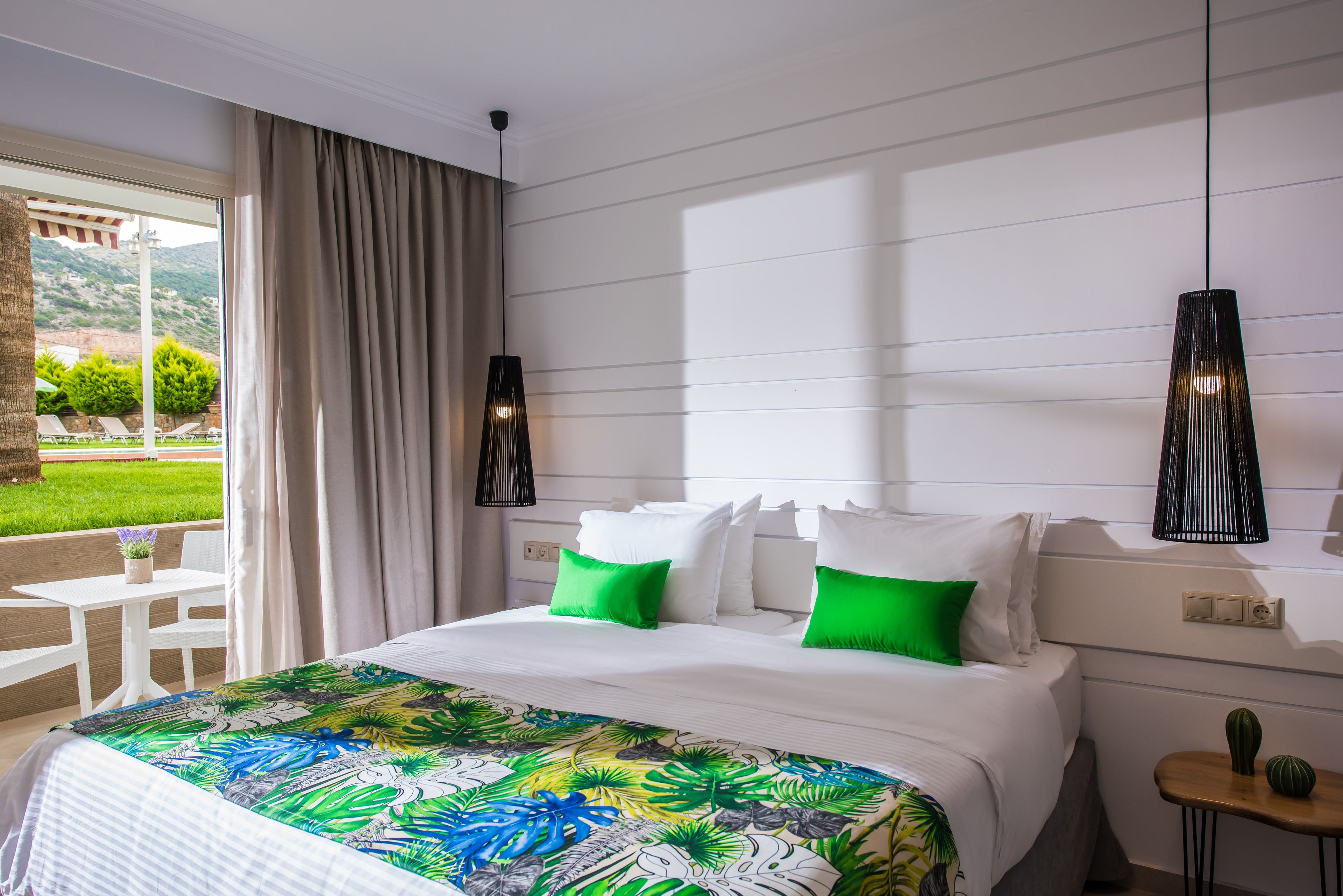 A hotel room with a bed, a nightstand, a table, and a view of a garden.