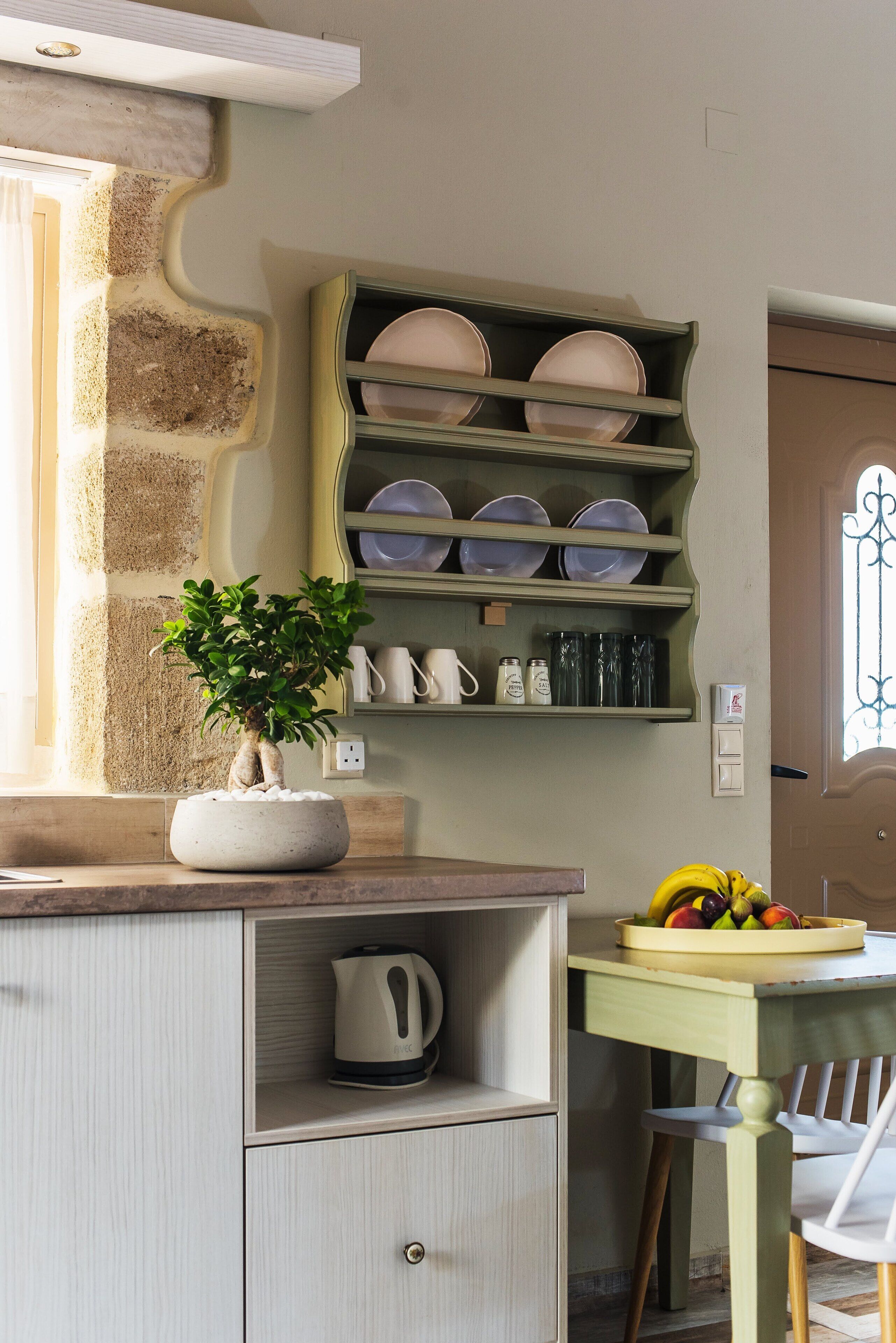 A kitchen with a built-in shelf holding plates and cups, a potted plant on the counter, and a fruit bowl on the table.