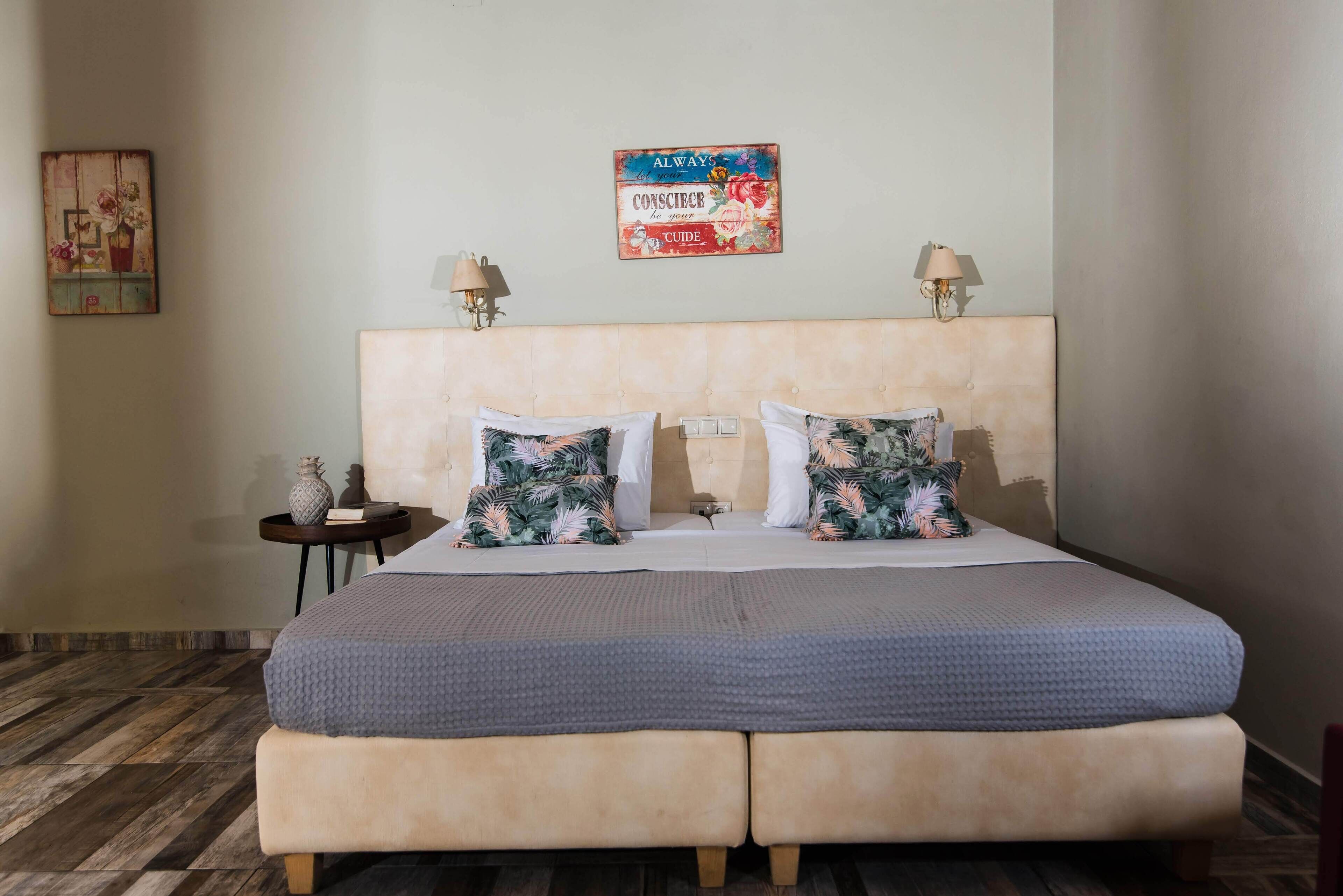 A double bed with a grey blanket, two matching pillows, a headboard with wall-mounted lamps, and a small table with a vase.