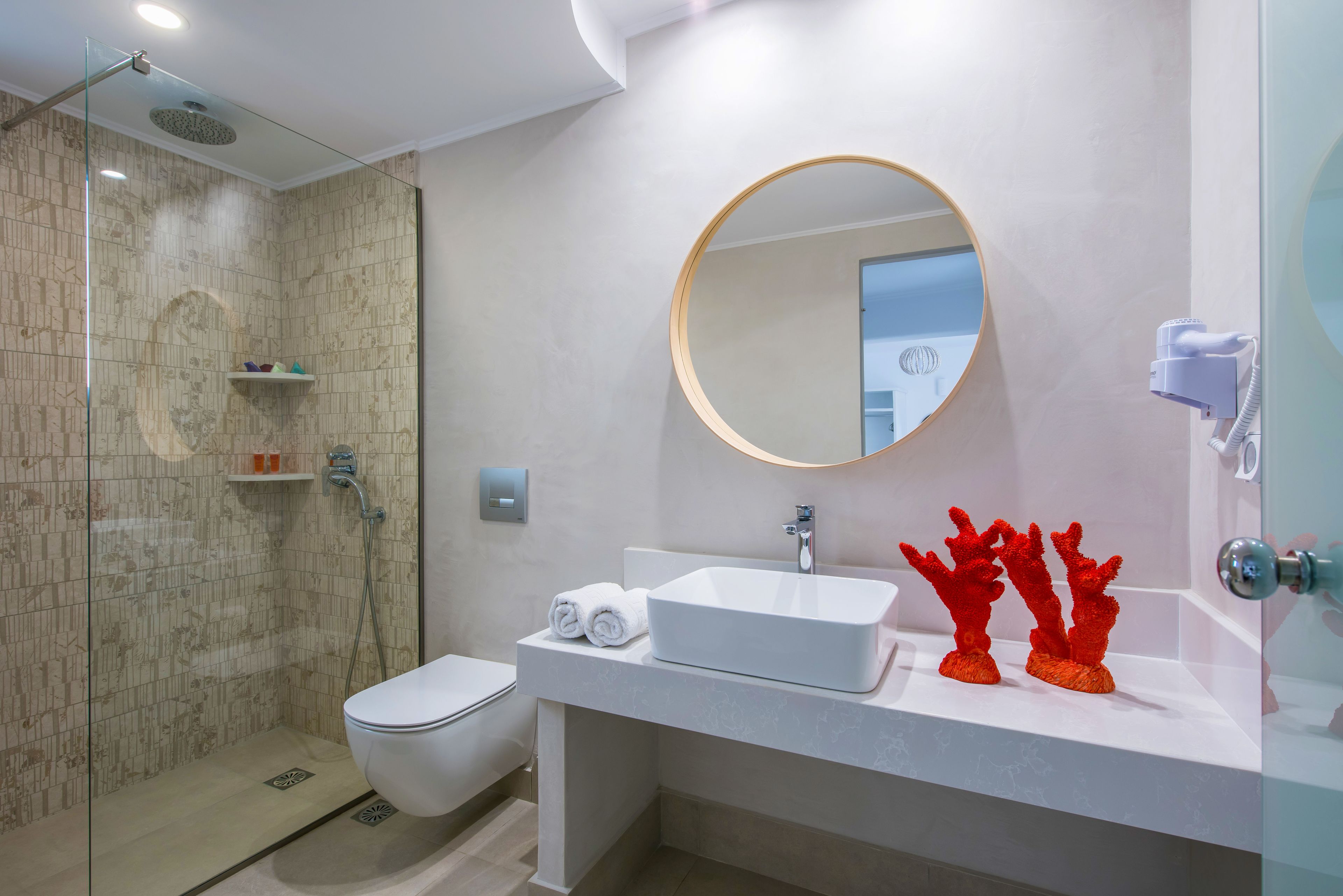 Un bagno moderno con doccia vetrata, un water, un lavandino con specchio tondo e sculture decorative a forma di corallo.
