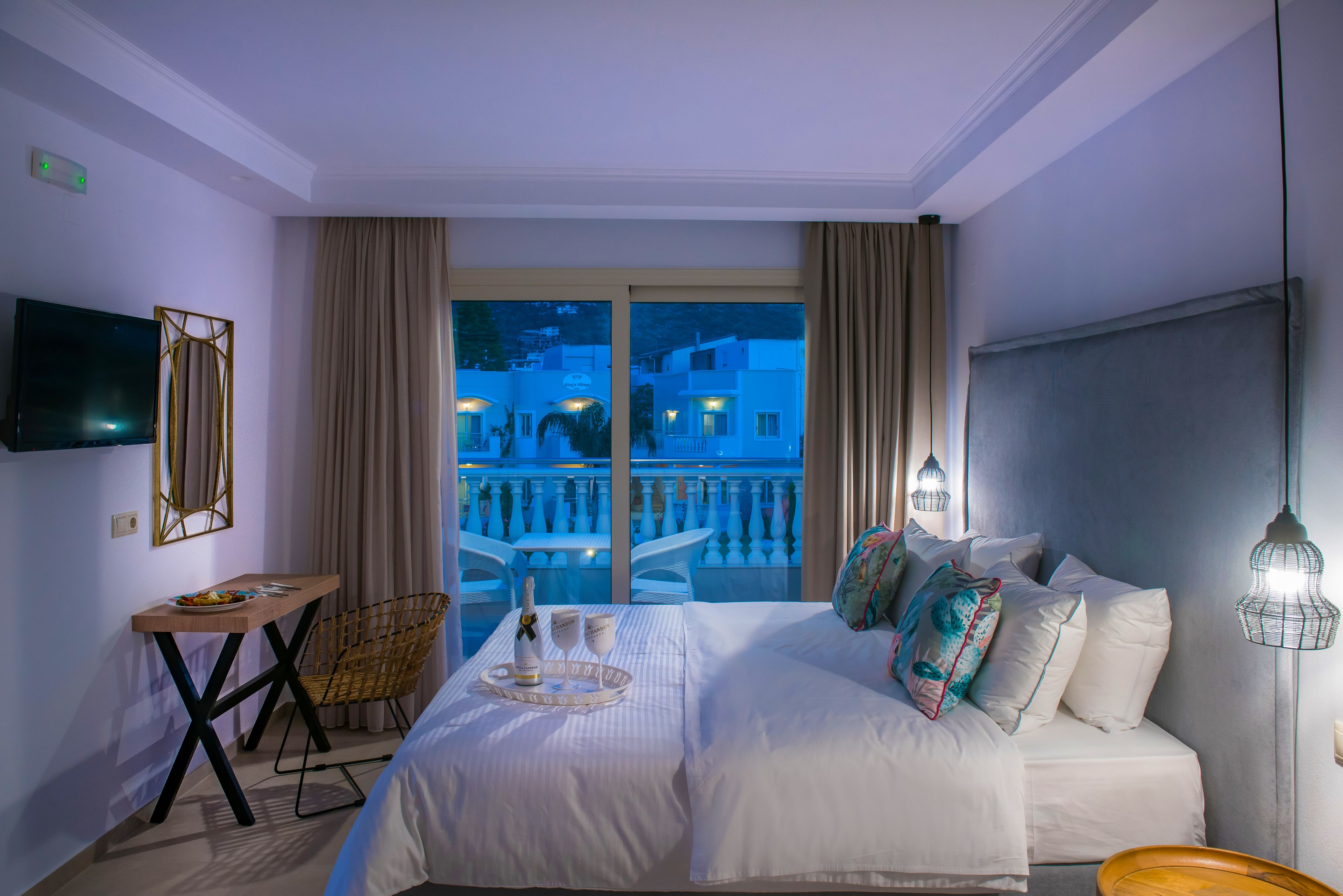 A hotel room with a bed, a desk, a chair, a TV, and a balcony with a view of buildings.