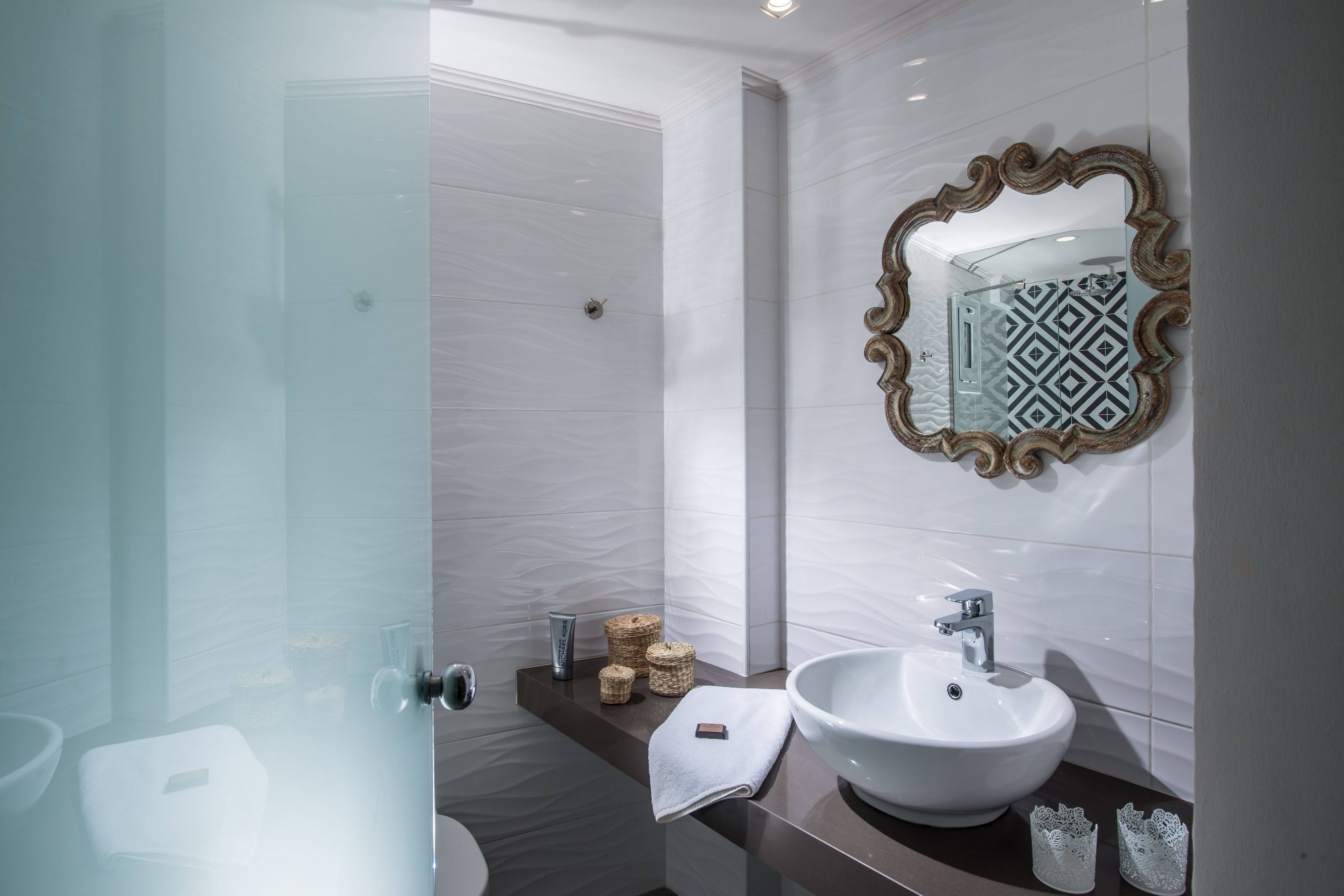 A modern bathroom with a glass shower enclosure, a white sink with a decorative mirror, and a small shelf with toiletries.