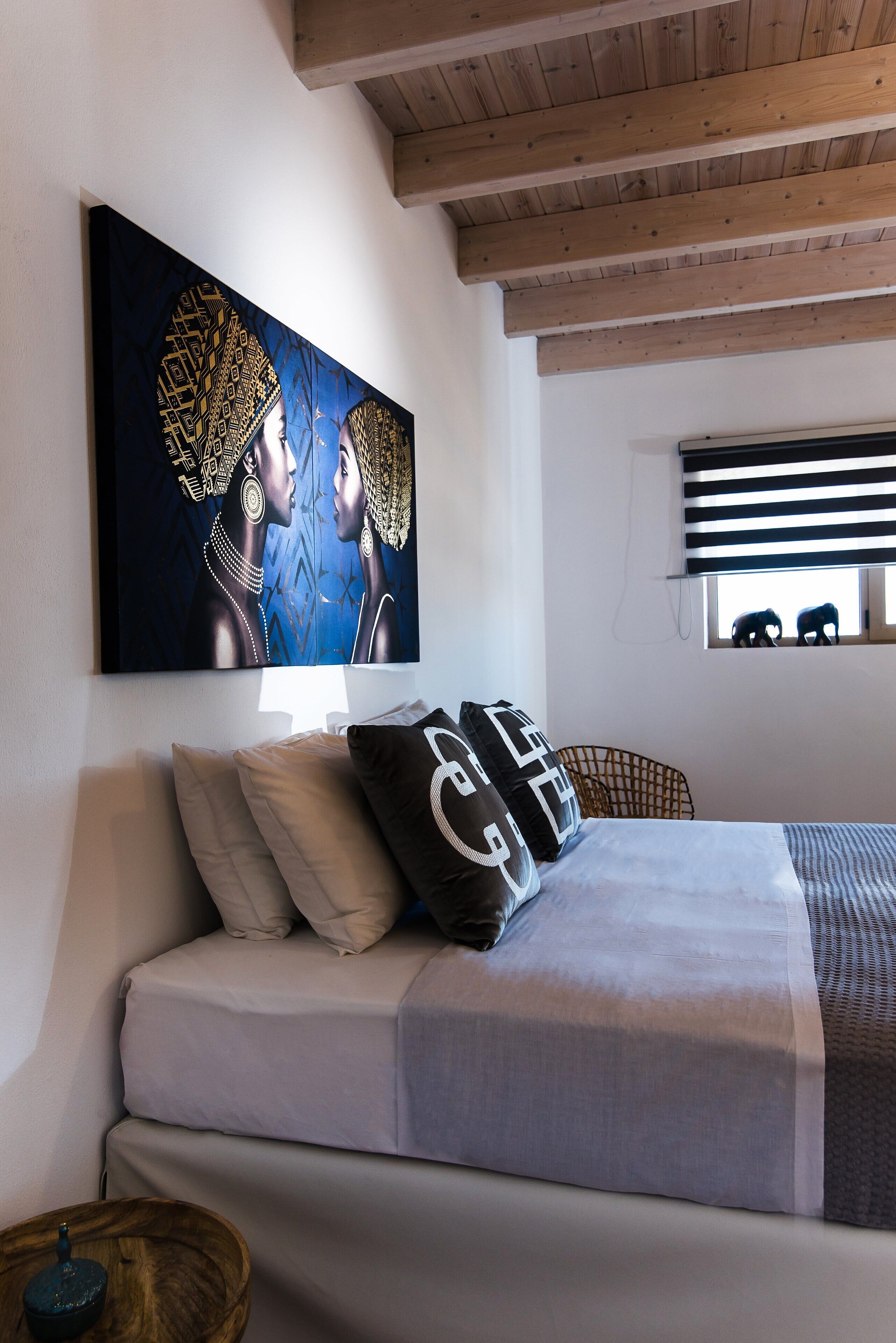 Una camera da letto con un letto, cuscini, un'opera d'arte appesa al muro, una finestra con persiane e un soffitto in legno.