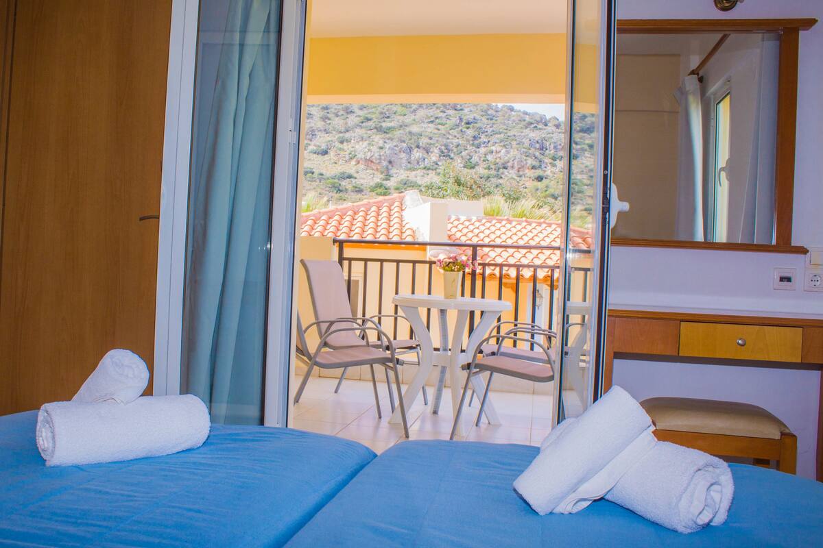 A hotel room with two beds, a balcony with a table and chairs, and a view of a hilly landscape.