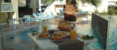 A poolside table with food and drinks, surrounded by lounge chairs and a palm tree.