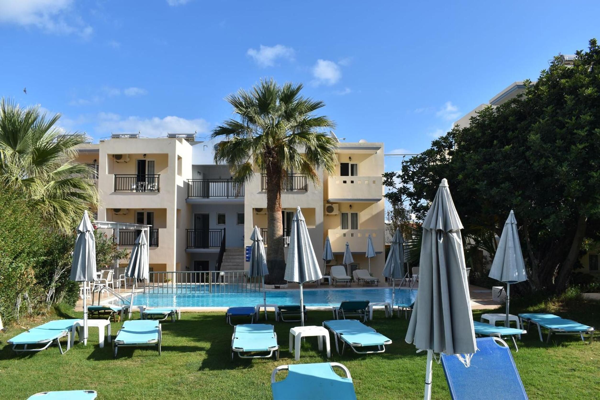 A pool area with lounge chairs and umbrellas, surrounded by a building with balconies and palm trees.