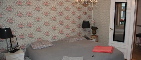 Un dormitorio con papel tapiz floral, una araña, una cama con cubrecama gris, dos mesitas de noche con lámparas y una puerta con espejo.