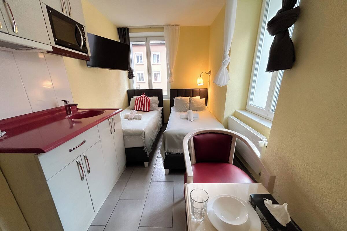 A compact hotel room with a kitchenette, two beds, a dining area, and a window with curtains.