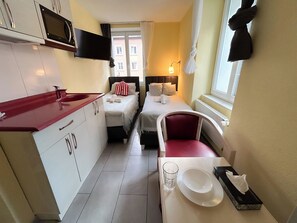 Una camera d'albergo compatta con angolo cottura, due letti, zona pranzo e finestra con tende.