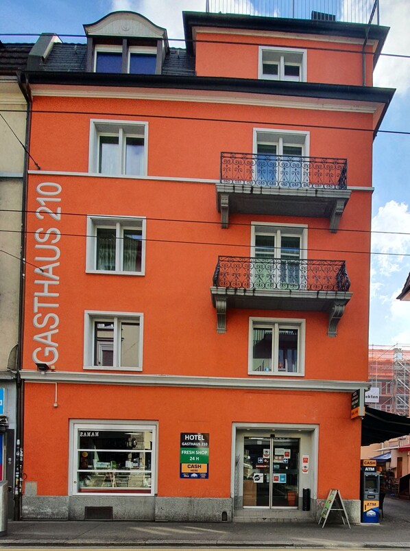 Un edificio a più piani con una facciata arancione brillante, un negozio al piano terra ben visibile e un cartello che indica un hotel e un negozio di prodotti freschi.