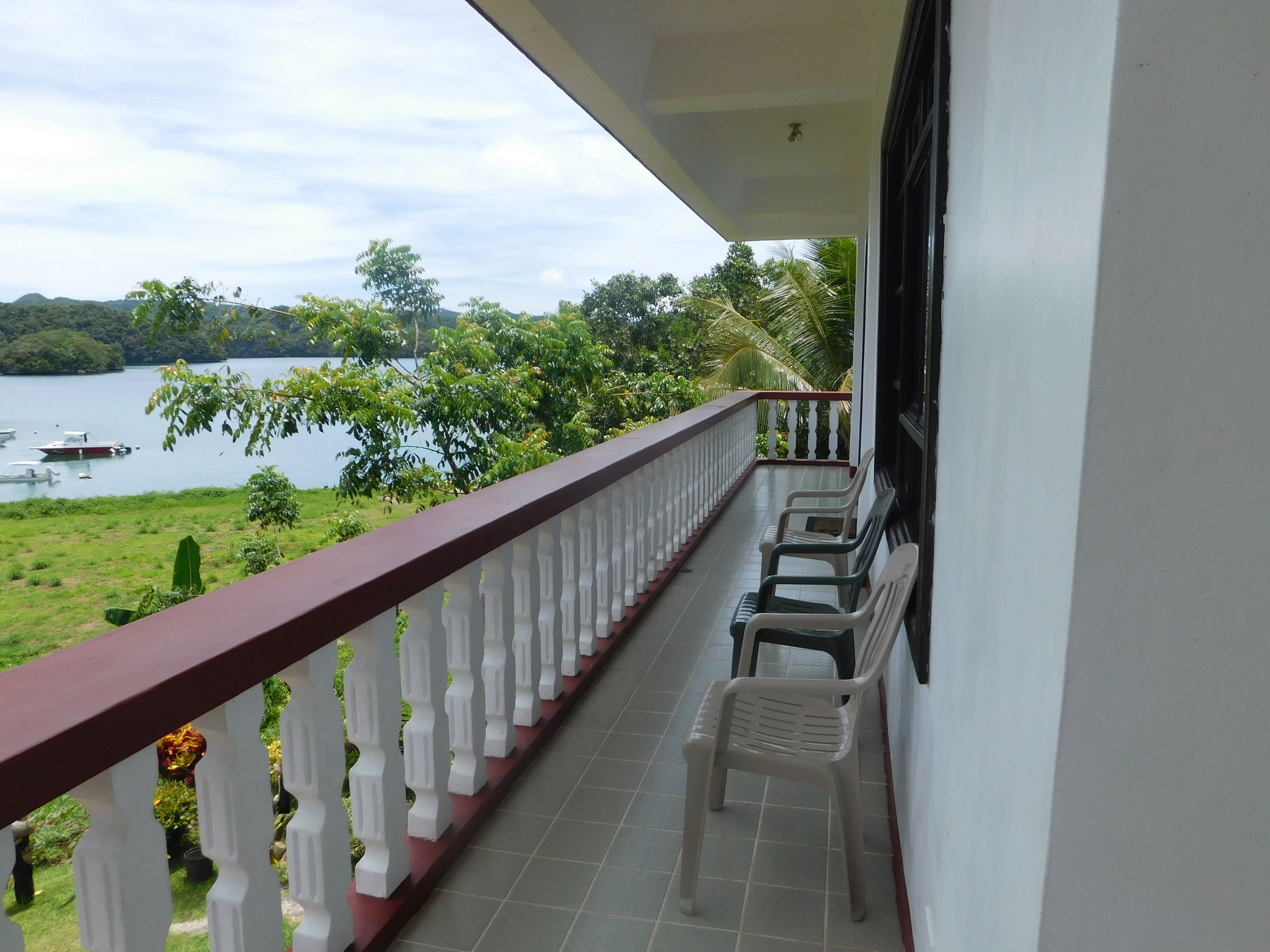 Uma varanda com guarda-corpo branco, algumas cadeiras de plástico e vista para um lago com barcos e vegetação.
