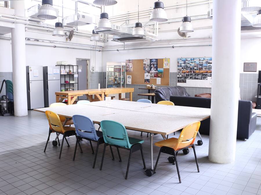 A spacious indoor area with a long table and colorful chairs, a bulletin board, and a refrigerator.