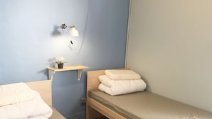 Quarto com duas camas, uma mesinha de cabeceira com luminária e uma pequena estante com planta em vaso.