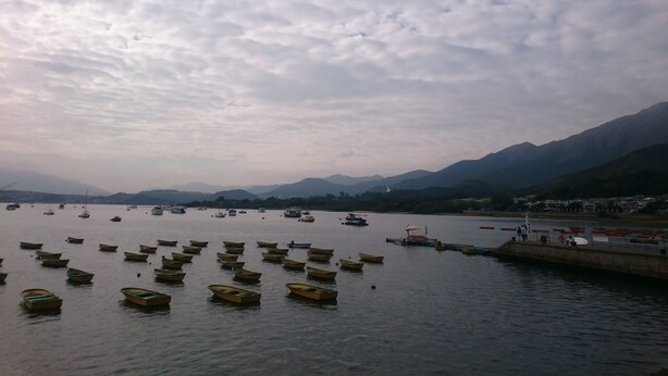 Um lago tranquilo com vários barcos pequenos ancorados ao primeiro plano, um cais com pessoas e montanhas ao fundo sob um céu nublado.