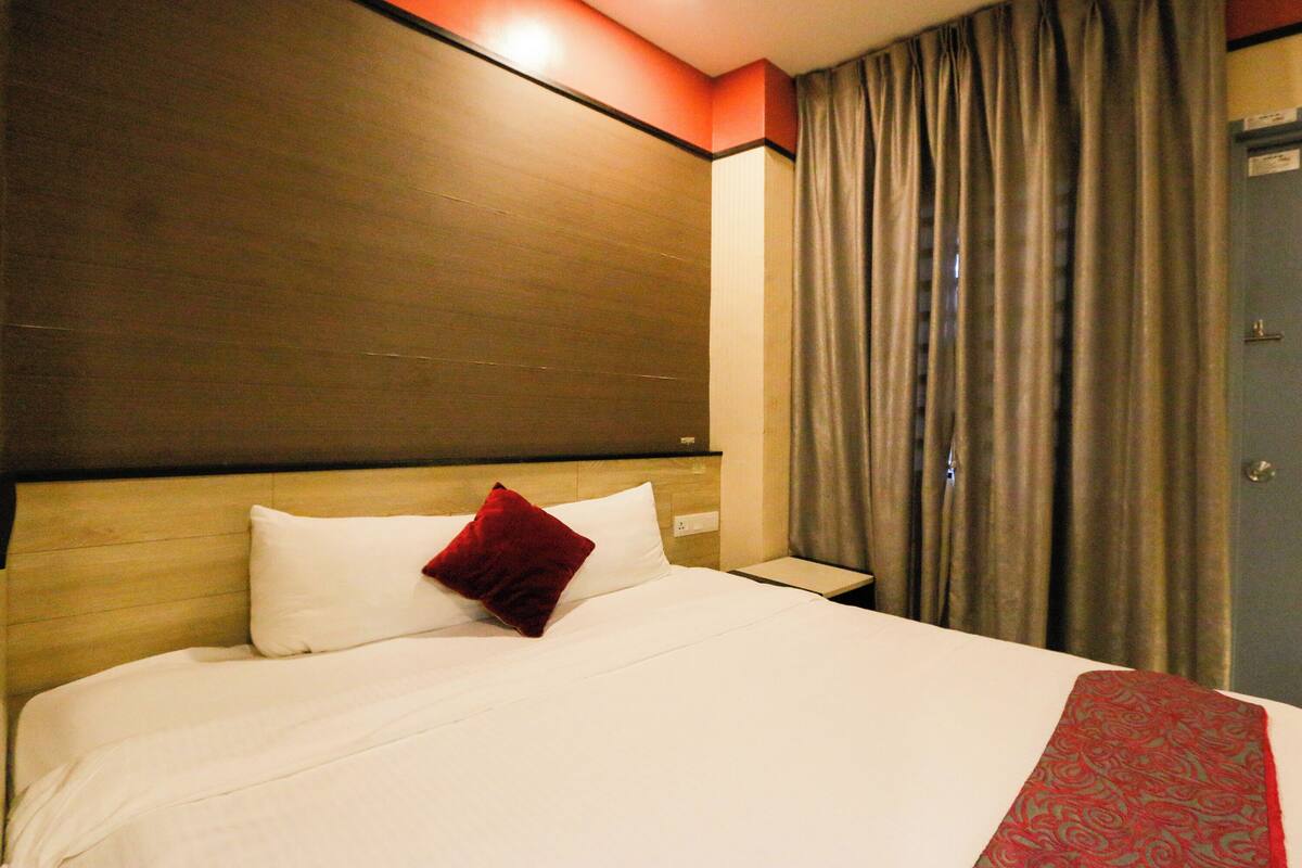 A hotel room with a large bed, a wooden headboard, a red pillow, a white pillow, a nightstand, and a door with a sign.