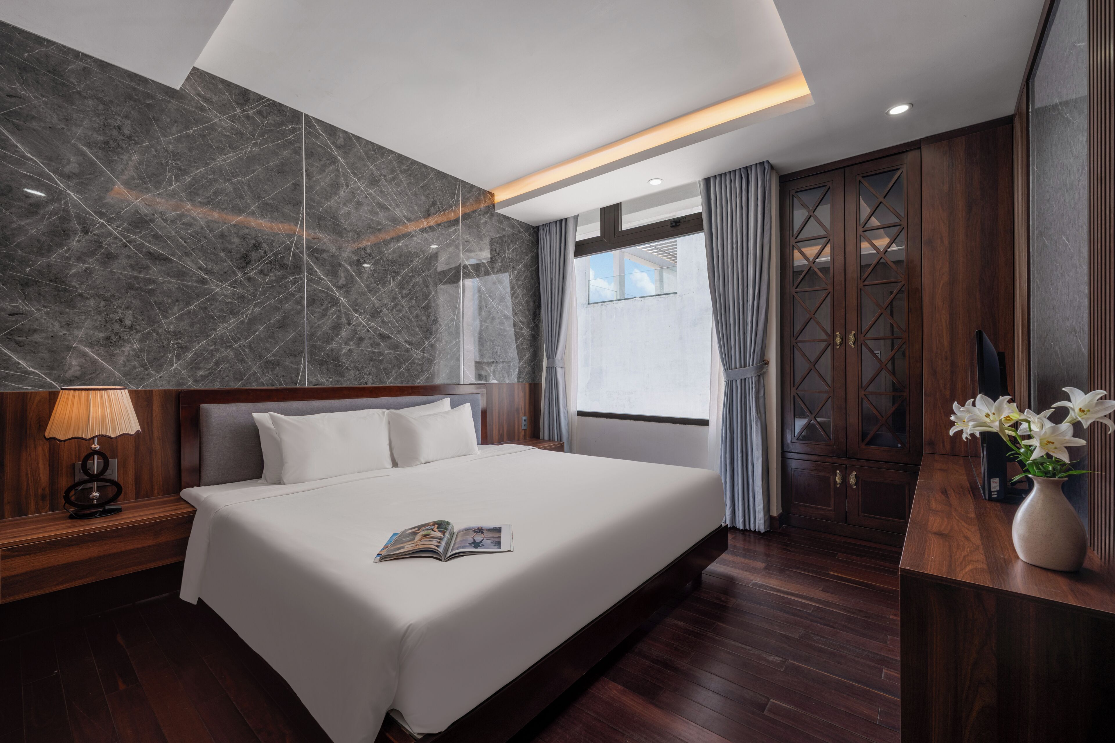 A modern hotel room with a large bed, wooden headboard, marble wall, and a view of the city.