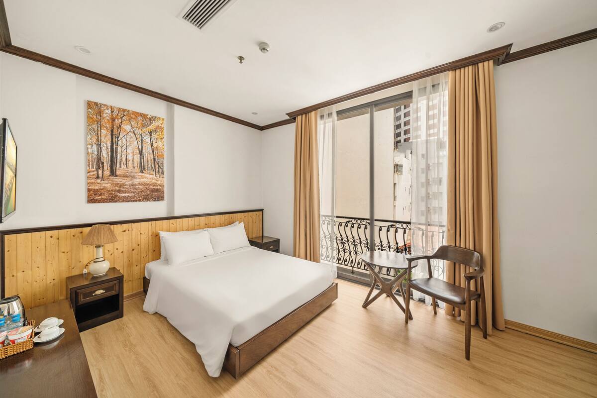 Ein Hotelzimmer mit einem großen Bett, einem kleinen Tisch, einem Sessel, einem Nachttisch, einem Gemälde und einem Balkon mit Vorhängen.