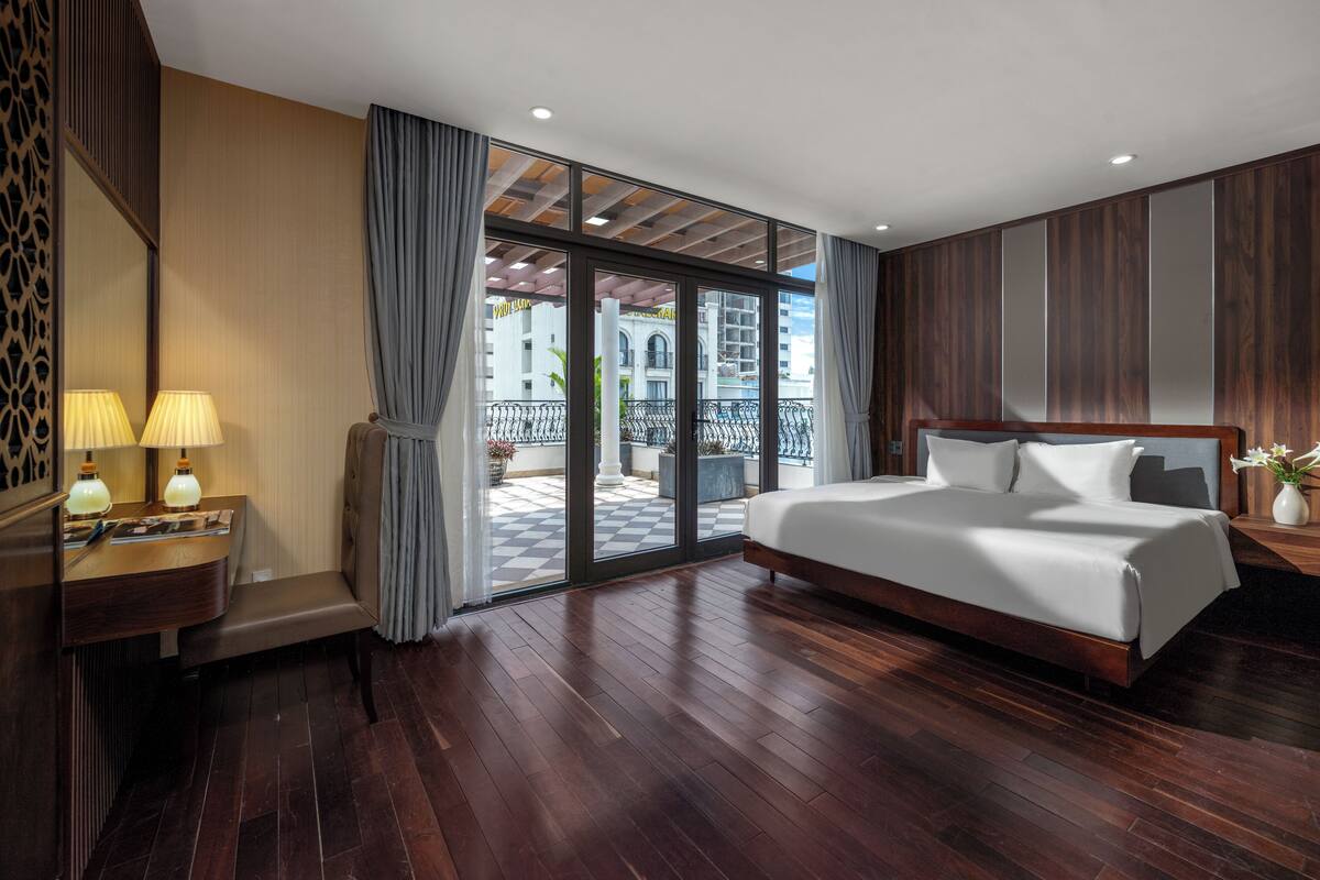 Ein Hotelzimmer mit einem großen Bett, einem Schreibtisch mit Lampen, einem Spiegel und Blick ins Freie.