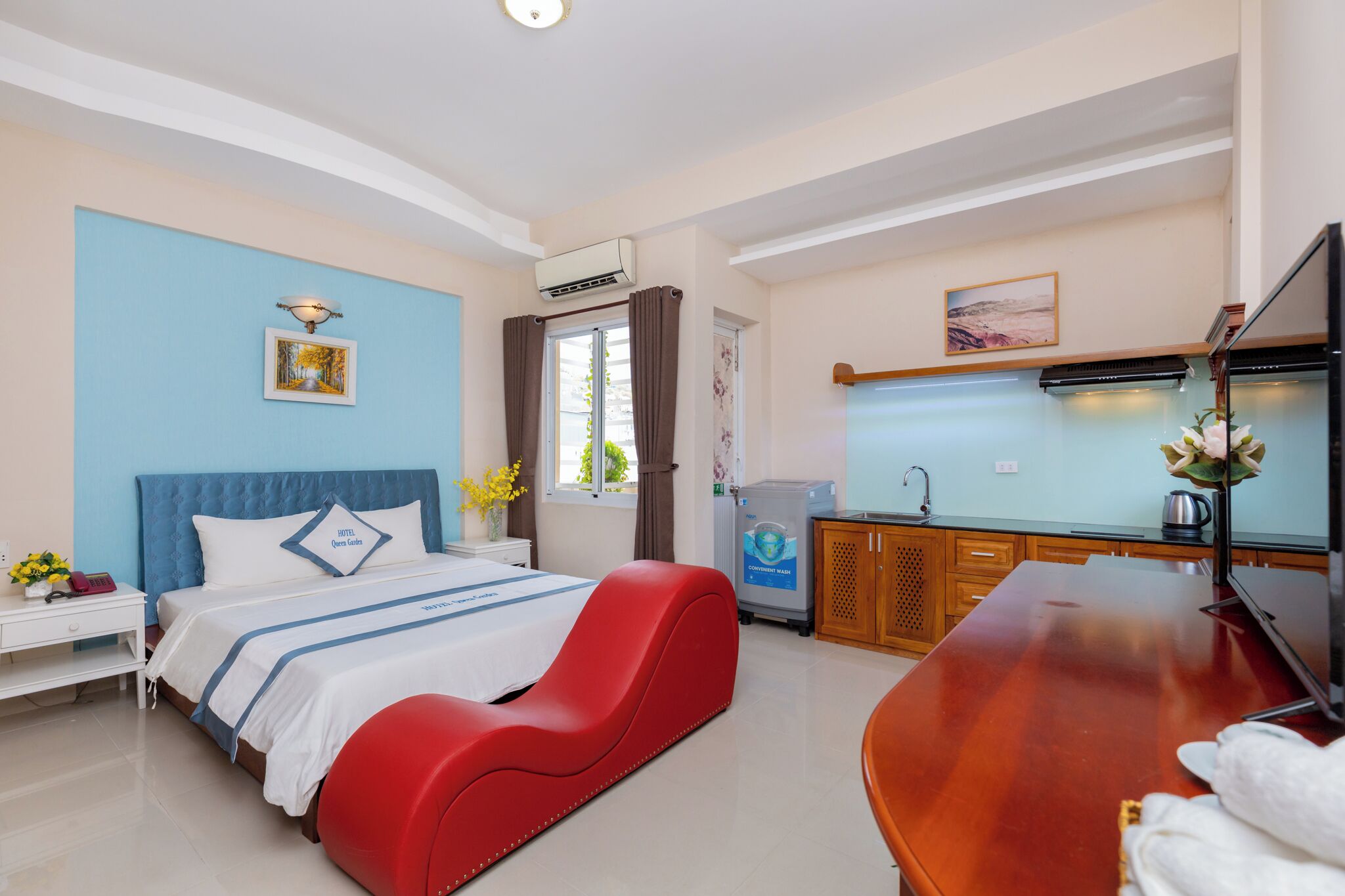 A hotel room with a bed, a red chair, a wooden table, a TV, a sink, and a window with curtains.