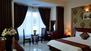 A hotel room with a bed, a desk, a chair, a small table with a vase of flowers, and a view of the outside through the window. - Queen Garden Hotel (Ho Chi Minh City)