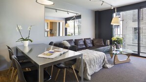 A modern living room with a glass dining table, black leather sofa, and a large window with blinds.