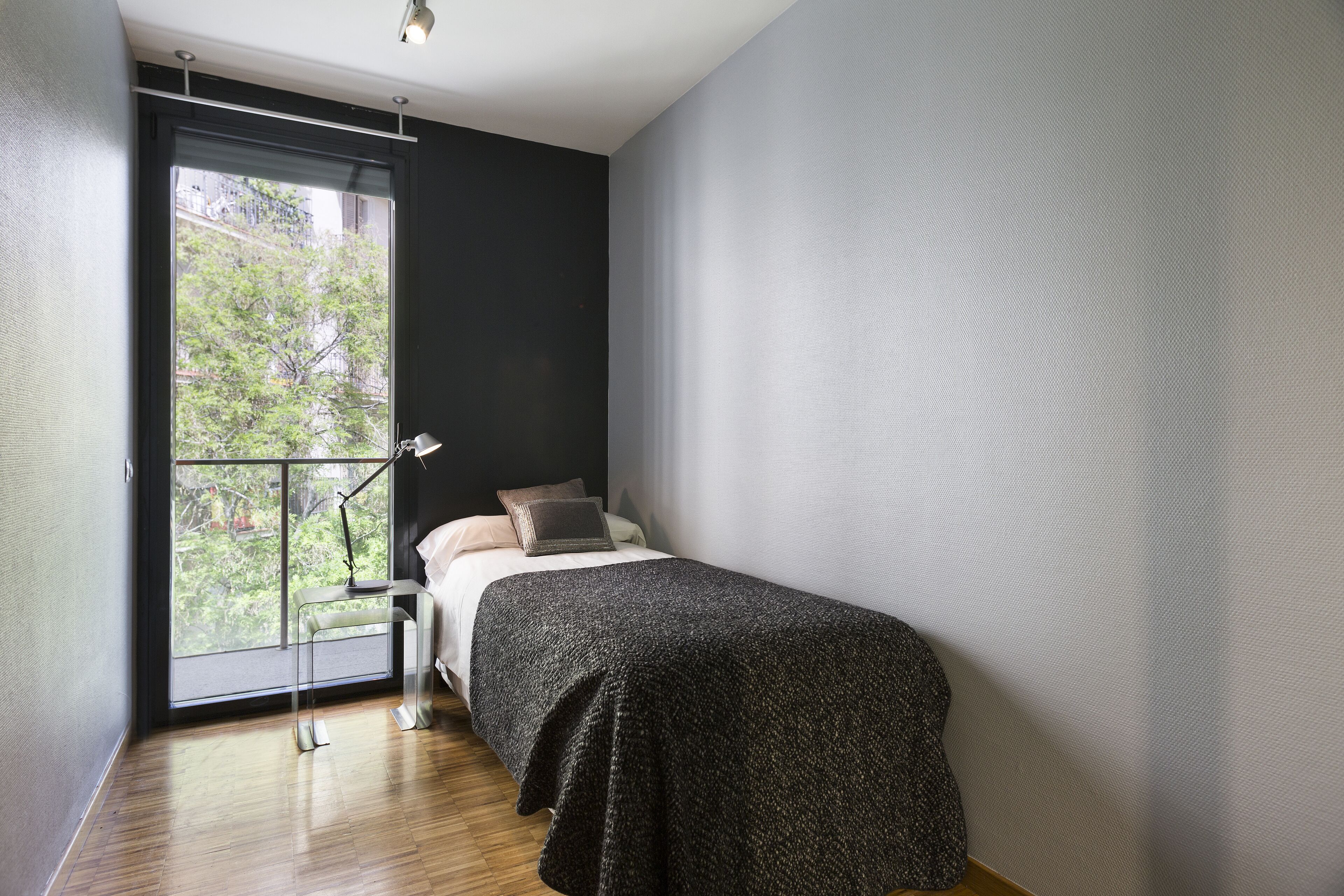 A bedroom with a large window, a bed, a nightstand, and a lamp.