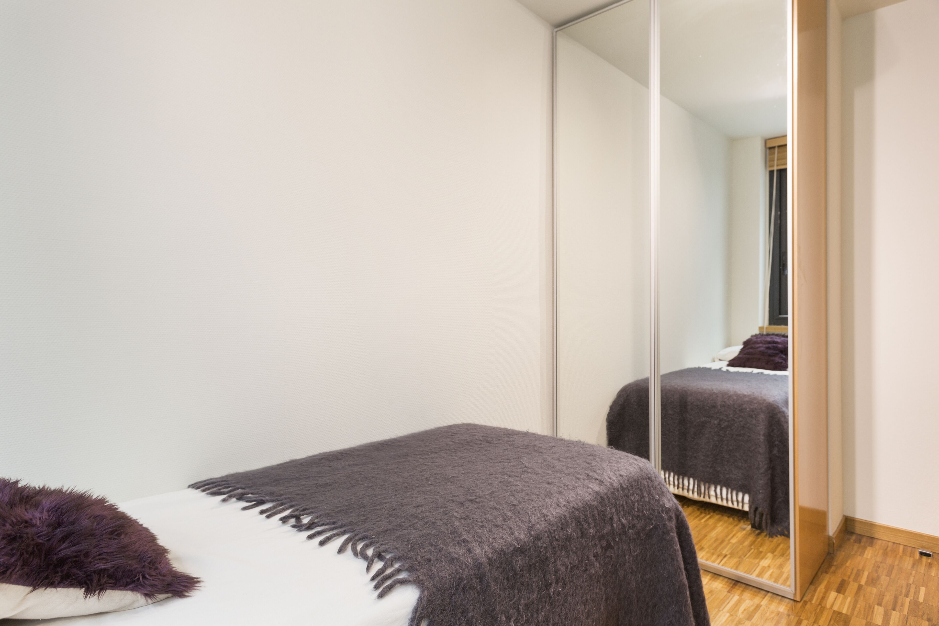 A bedroom with a large mirror, a bed with a dark blanket, and a small white bedside table with a purple pillow.