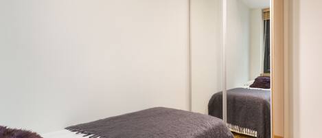 A bedroom with a large mirror, a bed with a dark blanket, and a small white bedside table with a purple pillow.