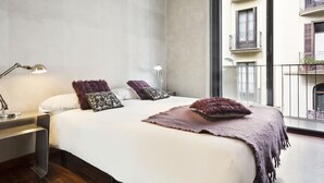 A modern bedroom with a large bed, a bedside table, a lamp, and a view of the city through a balcony.