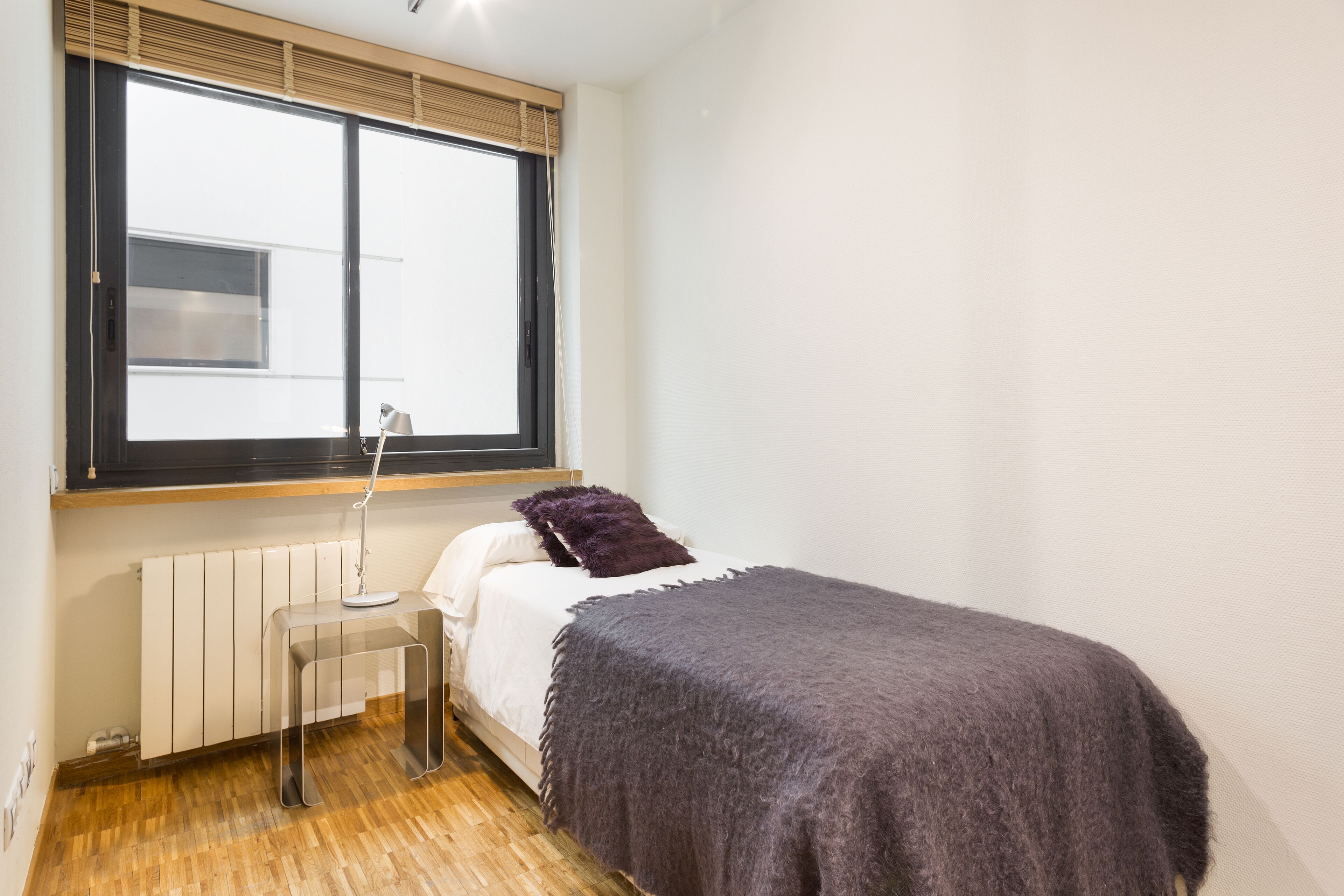 Une petite chambre à coucher, bien rangée, avec un lit simple, une table de chevet, une lampe et une fenêtre avec des stores.