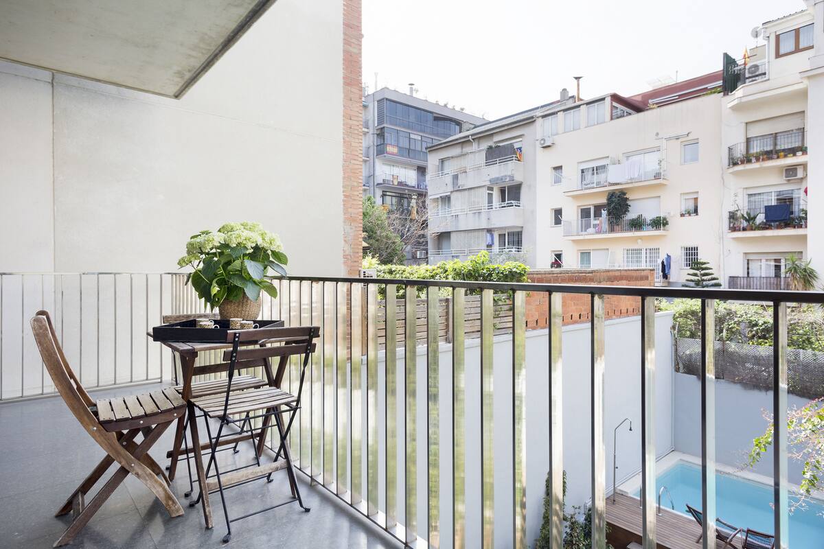 Un balcón con mesa y sillas, una planta en maceta y vistas a una piscina y edificios cercanos.