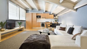 A modern living room with a sofa, ottoman, and a coffee table. There is a television on a stand, a kitchen area with cabinets and appliances, and a dining table with chairs.
