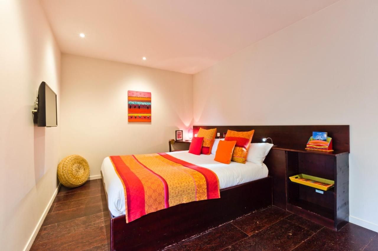 Un dormitorio con una cama, mesitas de noche, un televisor montado en la pared, una obra de arte colorida y un otoman tejido.