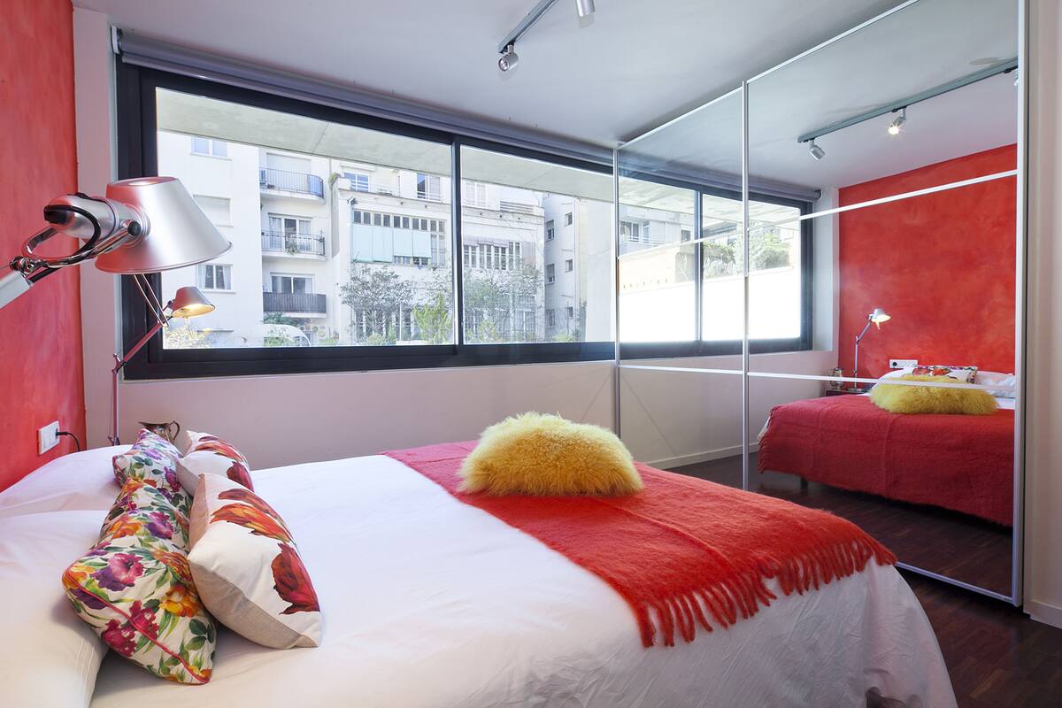 A modern hotel room with a large window, a bed with a red and white bedspread, a fluffy yellow pillow, and a mirror reflecting the room.