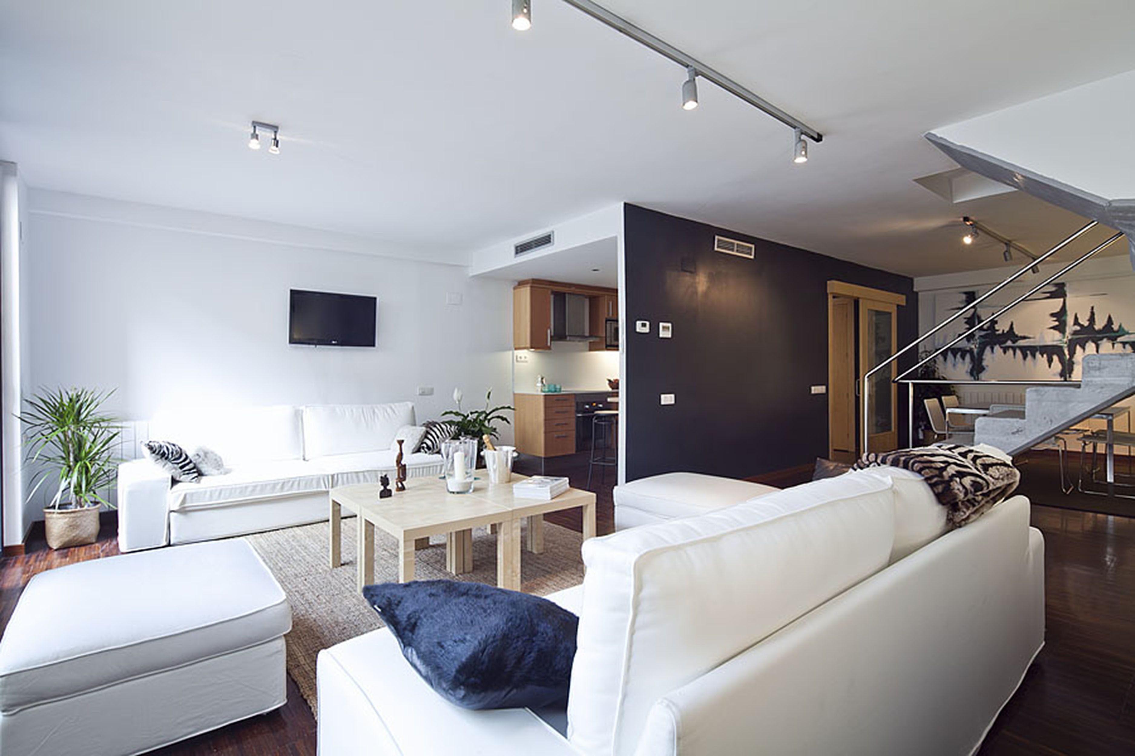 Una sala de estar moderna con un sofá blanco, una mesa de centro y un televisor montado en la pared. Se ve una escalera que sube a un nivel superior.