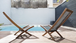Zwei Holzliegestühle an einem Pool mit einer weißen Wand und einem Balkon im Hintergrund.